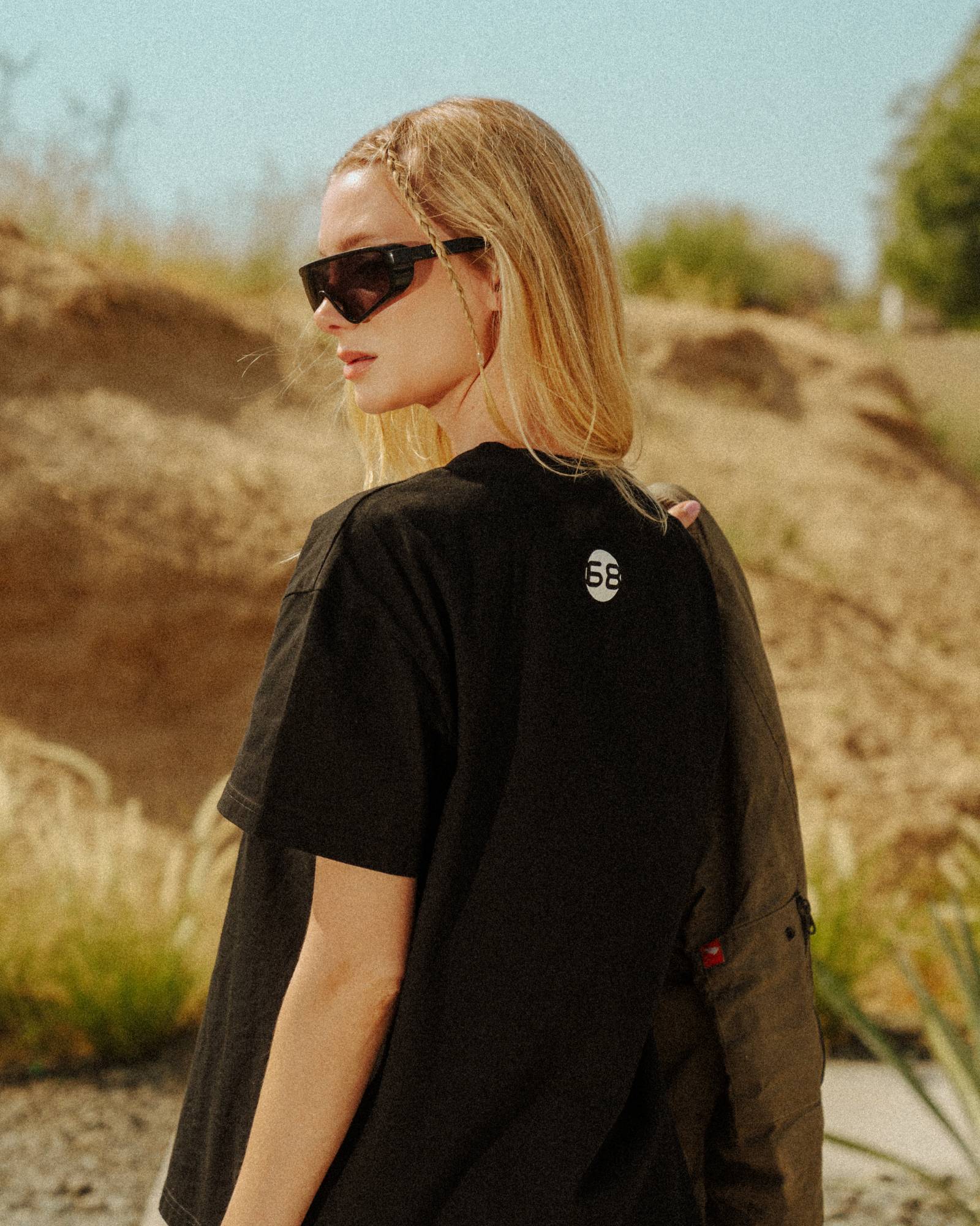 Figure seen from behind in an oversized black tee marked with the 68 roundel, sunlit desert scrub behind.