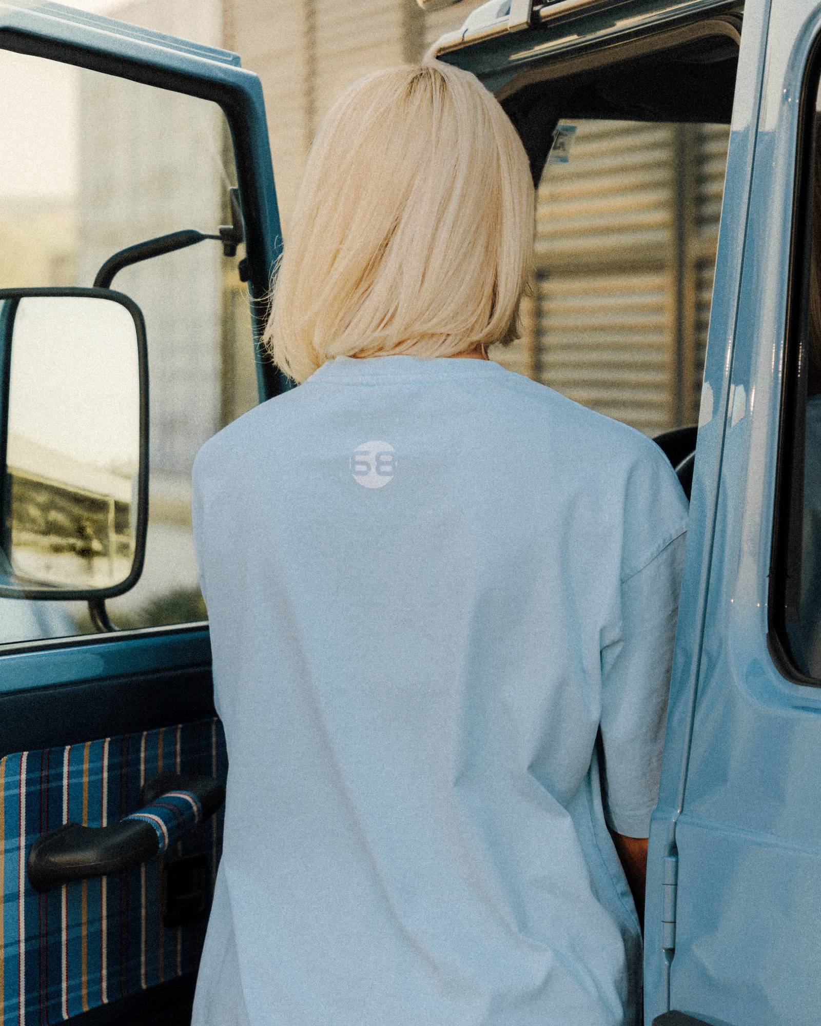 Figure from behind in a pale blue tee with the 68 roundel, platinum bob, stepping into a powder blue vintage truck.