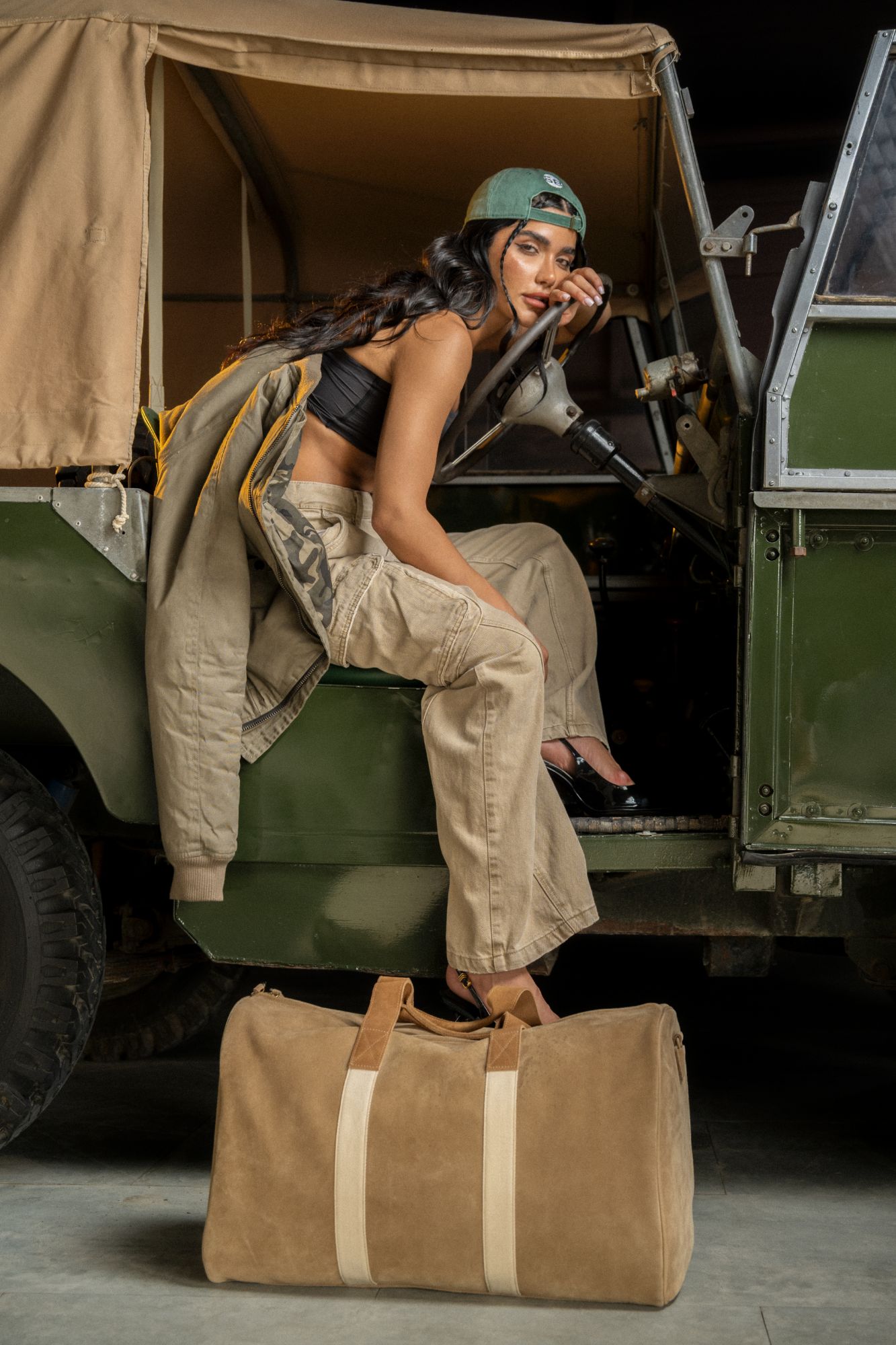 Figure perched in the doorway of a vintage green Land Rover, cap worn backwards, a tan suede duffel set down on the floor in the foreground.